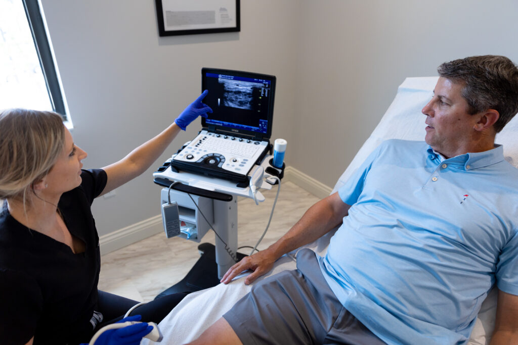 A female provider sits at the edge of a treatment bed, and speaks to a male patient about a vein ultrasound. The female provider discusses deep vein thrombosis treatment near Sarasota with the male patient.